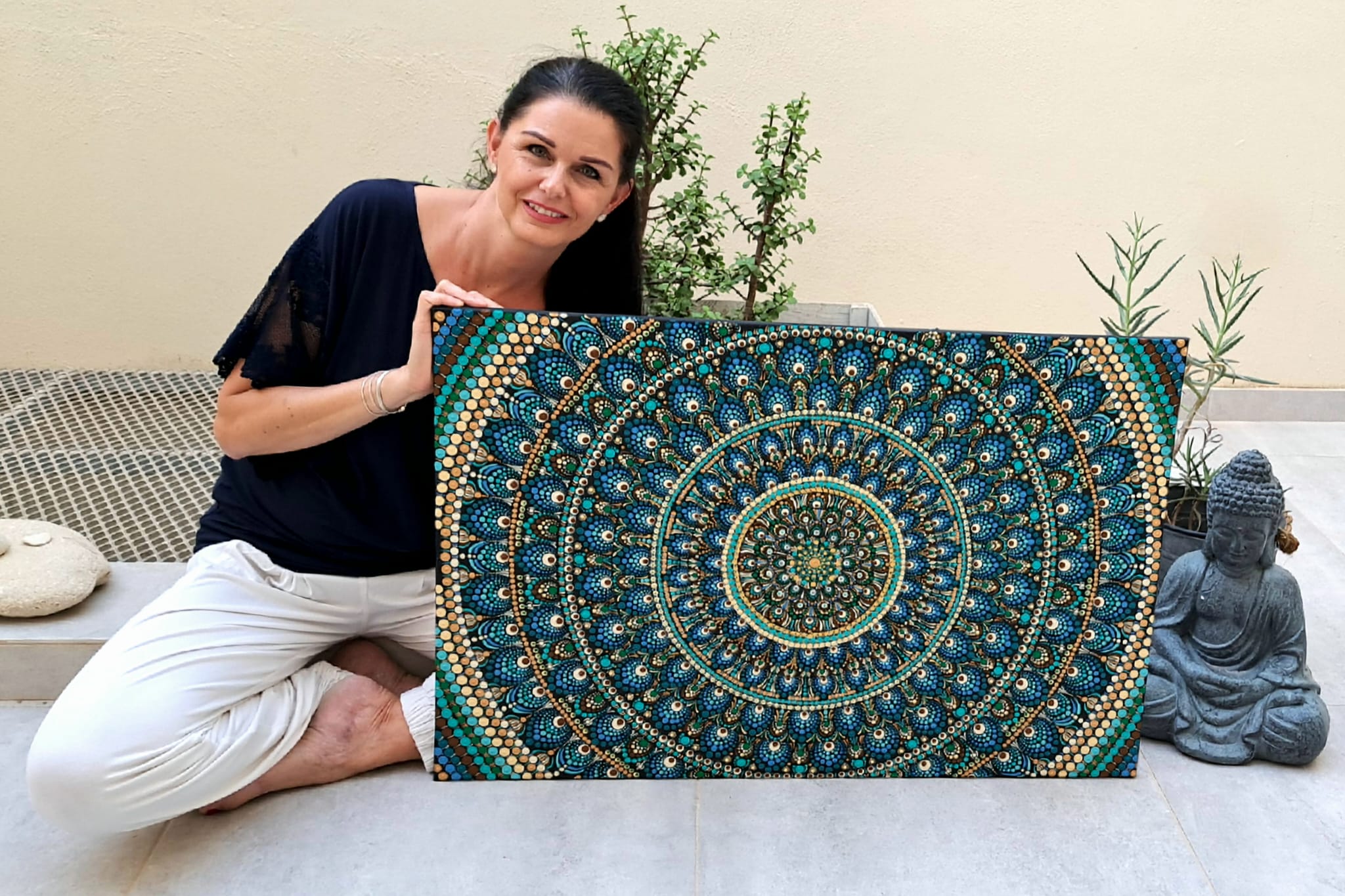 Woman holding hand-painted peacock mandala wall art in blue and gold tones, outdoor setting with Buddha statue